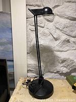 Full view of the black post modern tall table lamp with dome shade, positioned on a table against a stone wall background.