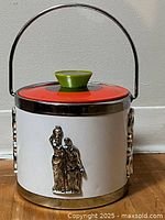 Front view showing cream-colored cylinder, red lid with green knob, metal handle, and bronze tone relief sculpture of two classical figures