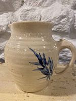 Front view of the cream pottery pitcher showing the body, handle, and flared top with blue floral decoration.