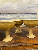 Three signed pottery goblets with yellow-brown glazed upper bowls and natural textured unglazed bases and stems, displayed on wooden surface against a painted backdrop.