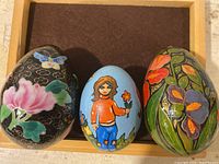 Set of three vintage hand-painted decorative eggs displayed on a brown felt background inside a wooden frame. Each egg has different folk art motifs: floral, butterflies, and a woman with a bird.