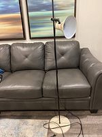 Full view of the mid-century modern tall floor lamp positioned in front of a grey sofa, showing its black slender pole, white conical metal shade and white circular base.