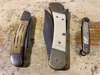 Three vintage folding pocket knives arranged on a wood surface, showing varying handle materials and blade sizes.