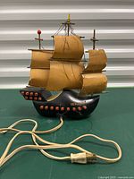 Front view of the black ship-shaped lamp with brown tattered sails, red cannon details, and attached electric cord with plug.