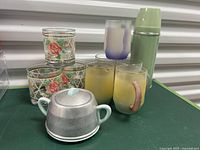 Five pastel mugs, three floral glasses, vintage sugar bowl with metal cover, and Acme thermos arranged on table.