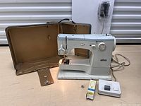 Full view of the Elna Super 62c Supermatic sewing machine with brown carrying case, foot pedal, and boxed light bulb on table.