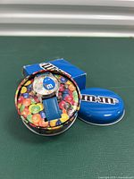 Blue M&M's character wristwatch with a blue strap inside a round collector's tin decorated with colorful M&M's candy imagery and M&M's logo on the lid.