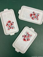 Three rectangular milk glass snack trays with red and pink floral Primrose design displayed on green background, showing overall lot contents and condition.
