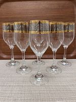 Set of six clear stemmed glasses with ornate gilded gold and silver rims featuring baroque design, arranged in a row on a beige cloth against wood background.