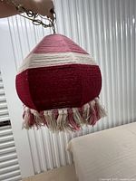 Handheld photo showing the vintage woven hanging lamp with multiple pink and white yarn stripes and yarn fringe on the edge, chain visible