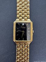 Front view showing square black dial with gold hour markers, hands and 'Caravelle by Bulova' branding, attached to multi-link gold tone metal bracelet.
