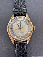 Front view of the watch displaying full watch face and gold-tone case with black strap.