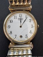 Close-up of the white dial featuring black Arabic numerals, minute markers, date window at 6 o'clock, and Seiko Quartz branding.