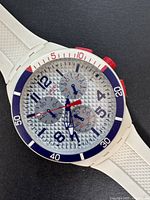 Closeup photo of watch face showing textured silver dial with blue numerals and red chronograph hands, white bezel with blue and red accents.