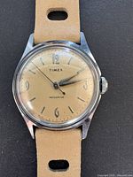 Close-up front view of vintage Timex men's watch with beige dial, stylized numerals, and beige leather strap.