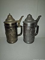 Two covered metal beer steins side by side showing detailed embossed designs and metal lids with thumb levers.