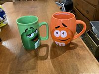 Shows front of green and orange M&M's character mugs side by side on wooden table.