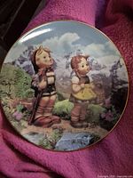 Front view of the M.J. Hummel 'Little Explorers' plate showing the detailed illustration of two children outdoors with mountains in the background, against a pink textile backdrop.