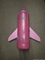 Top view of the pink Barbie airplane toy showing the closed body with wings. The airplane is pink with a small blue cockpit window, and has scratches and wear marks visible. A green sticker is seen on the body.