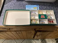 Open box displaying nine miniature ceramic teapots in individual compartments, each shaped like different buildings and a fire truck.