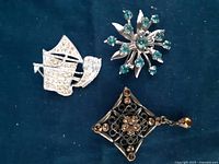 Three brooches shown together on a dark fabric background: a sailing ship with clear rhinestones, a silver-tone starburst with green gems, a black filigree square with amber gems and a teardrop.