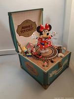 Angled view of vintage musical toy box with Minnie Mouse figurine on rotating platform and miniature train track with red toy train inside teal box with vintage decals.