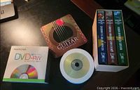 Group shot showing all items: Memorex DVD-RW box, Romantic Guitar tin, loose DVD spindle, boxed set of three VHS tapes