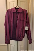 Front view of burgundy Functionals jacket on hanger showing full zip, flap pocket, elastic hem, attached retail tag