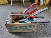 Lot overview inside Canada Dry crate containing mixed garden tools