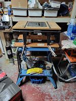 Overall lot showing router table top sitting on blue folding workbench with gloves and supplies below