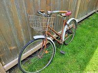 Full view of copper/bronze step-through bicycle with white fenders and basket against fence