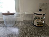 Four canisters lined up: frosted glass with wooden lid, two clear cylindrical jars with plastic lids, ceramic biscotti jar
