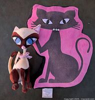 Plush Siamese cat toy sitting in front of a black cat pillow with purple background.