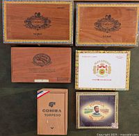 Six closed cigar boxes placed on green surface, showing brand logos and box dimensions.