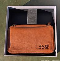 Top view of the closed brown leather cosmetic bag embossed with '360°' on front, placed in a white box with a dark interior.