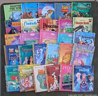 Photo shows 27 Disney children's storybooks laid out on a green surface, titles including Beauty and the Beast, Toy Story, Cinderella, Pinocchio, Sleeping Beauty, Tarzan, The Jungle Book, The Incredibles, and Frozen.
