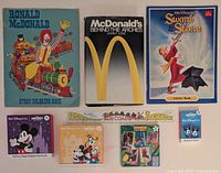 Overview of Ronald McDonald Story Coloring Book, McDonald's Behind the Arches hardcover, Sword in the Stone Activity Book, ruler coaster, and assorted Happy Meal toys including puzzles and memory game boxes.