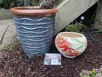 Photo of tall blue ceramic planter with wavy lines and small white rounded floral planter side by side outdoors on mulch under stairs.