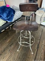 Front angle view of drafting chair showing dark wood seat and backrest and polished metal pedestal base with footrest ring.