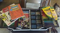 Opened black metal art suitcase showing compartments with colored pencils, oil pastels, and paint tubes