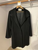 Black Fleurette cashmere wool 3/4 length coat, size 20, hanging on a wooden hanger in a white room.