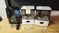 Two white Levoit ultrasonic humidifiers with digital displays and one black Honeywell humidifier with manual knob, cords included.