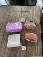 Five zippered pouches in metallic and patent finishes shown on a wood floor, along with metal necklace stand base