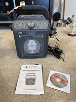 Front view of Singing Machine karaoke player, wired microphone plugged in, Party Tyme karaoke CD and instruction manual laid out on carpet.