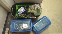 Top view of a green and black plastic bin filled with assorted Lego pieces including assembled figures and instruction booklets, alongside two sealed plastic boxes.