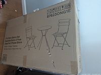 Sealed cardboard box showing printed illustration of 3-piece bistro set including one round table and two folding chairs in orange color.