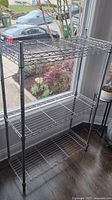 Full view of the silver metal wire shelving unit with 4 shelves, positioned in front of a window interior.