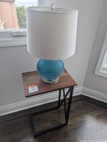Photo of the wood top and metal framed C shaped table with blue glass lamp and white shade placed on top near a window
