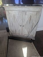 Front view of the whitewashed cabinet showing two decorative doors with diamond panel details and vertical grooves. Two dark knobs, cabinet placed on hardwood floor.