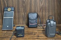 Four vintage cameras displayed front and center on wooden backdrop: Polaroid, Canon ELPH with case, Olympus with case, and Fuji TW-300 case